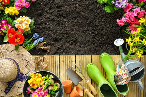The image depicts a section of a well-maintained garden with a wooden outdoor mat laid on the ground, bordered by various colorful flowering plants, including pink, yellow, and purple blossoms, along with a lush green hedge. In the foreground, there are gardening tools such as a small trowel, a hand rake, and an orange watering can, along with a pair of bright green rubber gardening boots and a garden hat decorated with a floral pattern. A black pot containing yellow and pink flowering plants is also visible on the mat. The background shows a patch of freshly turned soil, indicative of recent planting or soil preparation work. The overall scene captures a tidy, organized outdoor space suitable for gardening and lawn care activities, emphasizing the importance of garden maintenance in a typical UK suburban setting such as Brockley, to support local gardening services offered by Lawn Mowing Brockley.