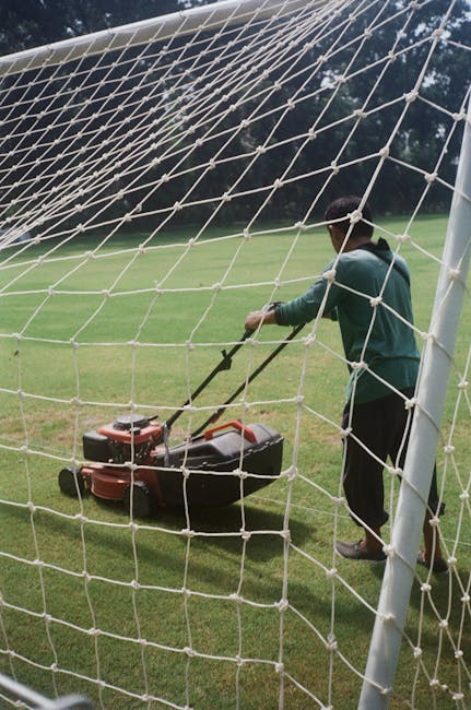 A young man operating a string trimmer in a well-maintained garden, surrounded by neatly trimmed evergreen hedges and lush green grass. The garden features a combination of different plants, with tall